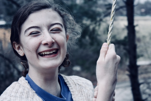 A young person is laughing joyfully while holding onto a rope. The background is slightly blurred with hints of greenery and a wooden structure.