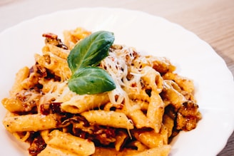 A white plate filled with creamy penne pasta, mixed with sun-dried tomatoes and garnished with fresh basil leaves. Grated cheese is sprinkled on top, adding a touch of elegance.