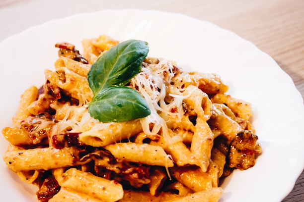 A white plate filled with creamy penne pasta, mixed with sun-dried tomatoes and garnished with fresh basil leaves. Grated cheese is sprinkled on top, adding a touch of elegance.
