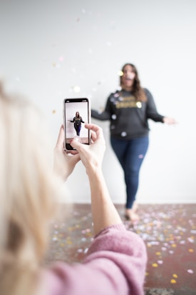 A person in the foreground, slightly out of focus, is holding a smartphone and capturing a photo of another person in the background. The second person is standing with arms open, surrounded by scattered confetti on the floor and in the air, giving a festive ambiance.