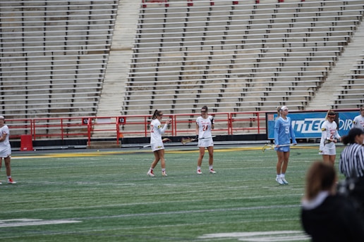 A group of athletes stands on a sports field, preparing for a game. The players wear uniforms, with some in white and one in light blue, and hold lacrosse sticks. The background shows empty stadium bleachers.
