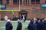 A group of people on a sports field, with some dressed in athletic gear, and others in casual clothing. A brick building labeled 'Gosset Football Team House' is in the background. One of the athletes appears to be aided by others, suggesting a possible injury.
