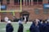 A group of people on a sports field, with some dressed in athletic gear, and others in casual clothing. A brick building labeled 'Gosset Football Team House' is in the background. One of the athletes appears to be aided by others, suggesting a possible injury.