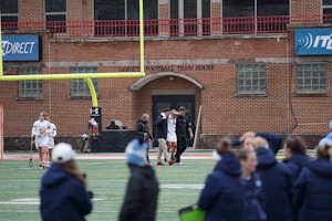 A group of people on a sports field, with some dressed in athletic gear, and others in casual clothing. A brick building labeled 'Gosset Football Team House' is in the background. One of the athletes appears to be aided by others, suggesting a possible injury.