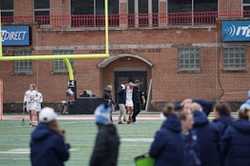 A group of people on a sports field, with some dressed in athletic gear, and others in casual clothing. A brick building labeled 'Gosset Football Team House' is in the background. One of the athletes appears to be aided by others, suggesting a possible injury.