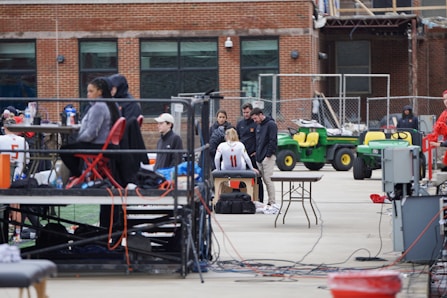 A group of people are gathered around an individual wearing a sports jersey with the number 11. They appear to be in a sports facility area, possibly a training or medical setup. There are tables, chairs, and equipment scattered around, with a brick building in the background. Some individuals are seated on a platform with electronics close by, suggesting a recording or monitoring activity. Utility vehicles and an orange barrier are present in the background.