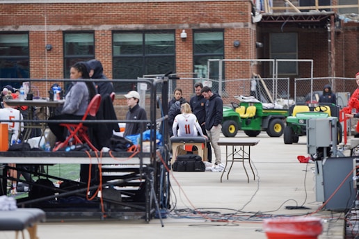 A group of people are gathered around an individual wearing a sports jersey with the number 11. They appear to be in a sports facility area, possibly a training or medical setup. There are tables, chairs, and equipment scattered around, with a brick building in the background. Some individuals are seated on a platform with electronics close by, suggesting a recording or monitoring activity. Utility vehicles and an orange barrier are present in the background.
