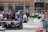 A group of people are gathered around an individual wearing a sports jersey with the number 11. They appear to be in a sports facility area, possibly a training or medical setup. There are tables, chairs, and equipment scattered around, with a brick building in the background. Some individuals are seated on a platform with electronics close by, suggesting a recording or monitoring activity. Utility vehicles and an orange barrier are present in the background.