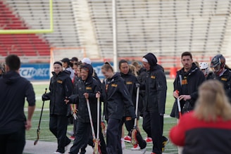A group of people wearing black jackets are gathered on a sports field. They are holding lacrosse sticks and some are conversing while others are looking towards the field. The background shows an empty stadium seating area.