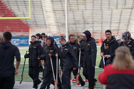 A group of people wearing black jackets are gathered on a sports field. They are holding lacrosse sticks and some are conversing while others are looking towards the field. The background shows an empty stadium seating area.