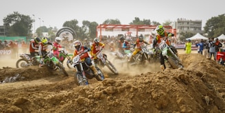 A group of motocross riders wearing colorful gear are racing on a dirt track, navigating a rough terrain with large mounds of dirt. Spectators watch from the sidelines, some capturing the event on cameras. The scene is filled with motion and dust, creating a sense of dynamic action.