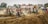 A group of motocross riders wearing colorful gear are racing on a dirt track, navigating a rough terrain with large mounds of dirt. Spectators watch from the sidelines, some capturing the event on cameras. The scene is filled with motion and dust, creating a sense of dynamic action.