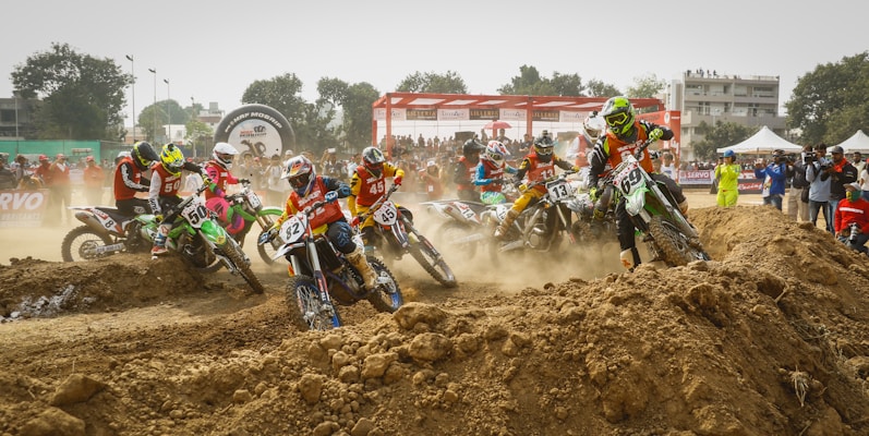 A group of motocross riders wearing colorful gear are racing on a dirt track, navigating a rough terrain with large mounds of dirt. Spectators watch from the sidelines, some capturing the event on cameras. The scene is filled with motion and dust, creating a sense of dynamic action.