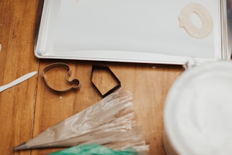 On a wooden surface, there are several baking tools including cookie cutters, a piping bag, and a metal baking tray. The cookie cutters are shaped like a teapot and a hexagon, and a partially cut piece of dough is also visible.