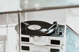 A vintage-style record player with an open lid, featuring a black vinyl record spinning on the turntable. It sits on a reflective glass surface, with soft natural lighting creating a serene atmosphere.