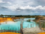 A scenic view of a vibrant turquoise lake surrounded by wooden pathways. The landscape features yellow-orange sandy patches and green foliage in the background. A small red flag stands atop a hill, with cloudy skies creating a tranquil, expansive atmosphere.