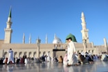 A large mosque with multiple domes and minarets against a clear blue sky. Numerous people dressed in traditional clothing are gathered outside, some sitting or walking on a polished stone surface.