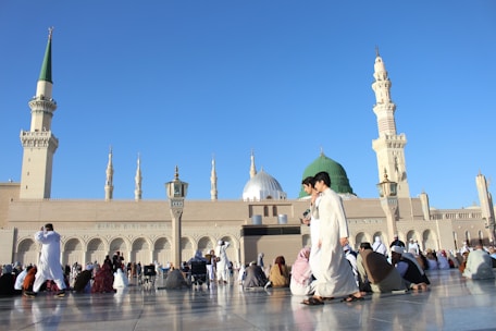 A large mosque with multiple domes and minarets against a clear blue sky. Numerous people dressed in traditional clothing are gathered outside, some sitting or walking on a polished stone surface.