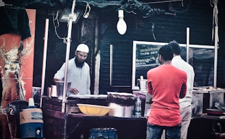 A street vendor is preparing food at a small outdoor stall. The vendor, wearing a white cap and shirt, is focused on his task. Two customers are standing in front of the stall, one in a red shirt and the other in a white shirt. A Coca-Cola advertisement is visible on the left. Various cooking utensils and a water container are present on and around the stall.