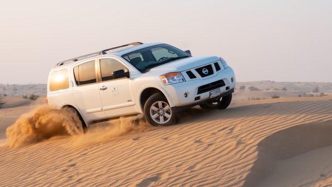 A white SUV drives over sandy desert dunes, lifting dust as it maneuvers. The background shows a vast expanse of desert with gentle hills under a clear sky.
