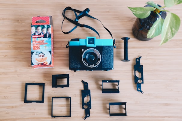 A vintage-style camera with a turquoise top and a black body is placed on a wooden surface. Surrounding the camera are various black plastic components, likely parts of the camera. To the left, there is a box of Lomography color negative film featuring bright packaging with portraits and a large orange fruit picture. A small plant in a glass vase is positioned on the right side of the composition.