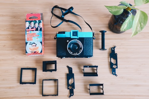 A vintage-style camera with a turquoise top and a black body is placed on a wooden surface. Surrounding the camera are various black plastic components, likely parts of the camera. To the left, there is a box of Lomography color negative film featuring bright packaging with portraits and a large orange fruit picture. A small plant in a glass vase is positioned on the right side of the composition.