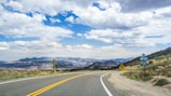 A scenic highway curves through a mountainous landscape with road signs visible on the right. The sky is partially cloudy, allowing glimpses of blue, and the surrounding terrain consists of rolling hills and distant mountains.