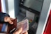 A person is holding a wallet open with several credit cards visible inside, along with a stack of paper currency partially pulled out. In the background, an ATM keypad and screen are visible, indicating a transaction in progress.