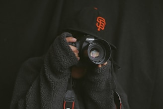 A person wearing a textured dark sweater and a baseball cap is holding a Canon camera up to their face, as if taking a photograph. The cap features an orange logo, and the background is dark, creating a dramatic atmosphere.