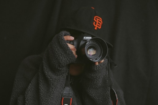 A person wearing a textured dark sweater and a baseball cap is holding a Canon camera up to their face, as if taking a photograph. The cap features an orange logo, and the background is dark, creating a dramatic atmosphere.