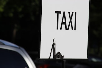 A sign with the word 'TAXI' written in bold black letters on a white background. Below the word, there is an illustration of a person sitting with their knees up, depicted in a minimalist black silhouette style. Part of a vehicle is partially visible in the foreground on the left side of the image.