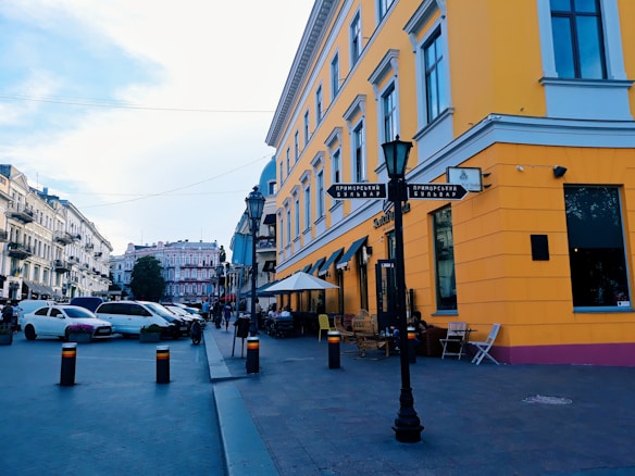A vibrant yellow building is situated on a street corner, with a series of outdoor tables and chairs under awnings. Several cars are parked on the street, and pedestrians can be seen walking along the sidewalk. The architecture is European-style, with detailed facades and multiple stories. Nearby buildings are painted in lighter shades, and the sky is clear with a touch of clouds.