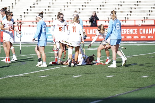 Lacrosse players on a field surround and attend to a player lying on the ground. The players are wearing sports uniforms, with some in white and others in light blue. The background shows bleachers and a red banner with white text advertising various championships.