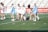 Lacrosse players on a field surround and attend to a player lying on the ground. The players are wearing sports uniforms, with some in white and others in light blue. The background shows bleachers and a red banner with white text advertising various championships.
