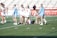 Lacrosse players on a field surround and attend to a player lying on the ground. The players are wearing sports uniforms, with some in white and others in light blue. The background shows bleachers and a red banner with white text advertising various championships.