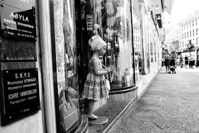 A mannequin resembling a child is dressed in patterned clothing and positioned in front of a store window on a busy urban street. Several informational signs in French are displayed nearby. People are walking along the street, some with strollers, and classic European-style buildings are visible in the background.