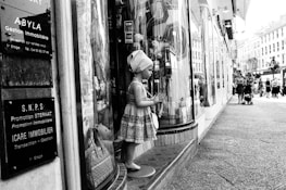A mannequin resembling a child is dressed in patterned clothing and positioned in front of a store window on a busy urban street. Several informational signs in French are displayed nearby. People are walking along the street, some with strollers, and classic European-style buildings are visible in the background.