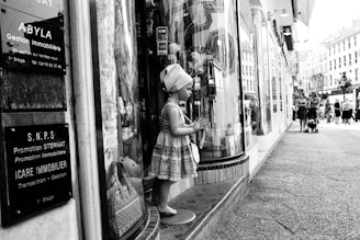 A mannequin resembling a child is dressed in patterned clothing and positioned in front of a store window on a busy urban street. Several informational signs in French are displayed nearby. People are walking along the street, some with strollers, and classic European-style buildings are visible in the background.