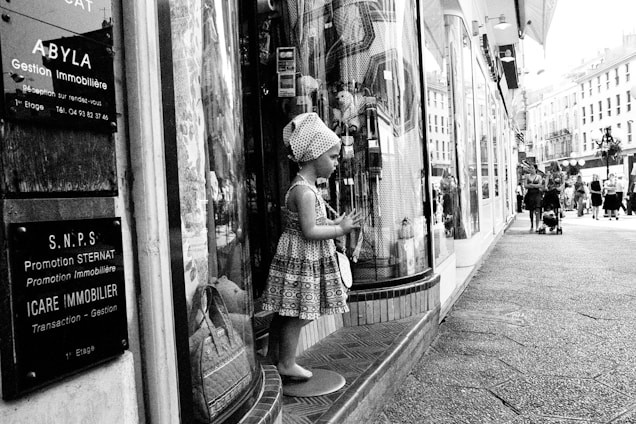 A mannequin resembling a child is dressed in patterned clothing and positioned in front of a store window on a busy urban street. Several informational signs in French are displayed nearby. People are walking along the street, some with strollers, and classic European-style buildings are visible in the background.