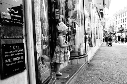 A mannequin resembling a child is dressed in patterned clothing and positioned in front of a store window on a busy urban street. Several informational signs in French are displayed nearby. People are walking along the street, some with strollers, and classic European-style buildings are visible in the background.