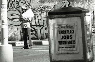 A man stands in an urban environment holding a large paper or cardboard object. The background features a graffiti-covered wall with bold lettering and numbers. In the foreground, an advertisement board displays a sign reading 'WORKPLACE JOBS WEDNESDAYS' in large letters. The scene is in black and white, creating a gritty, urban aesthetic.