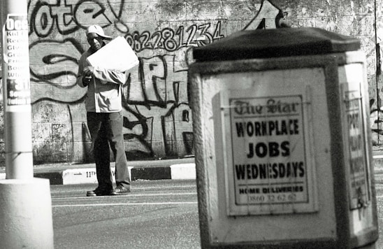 A man stands in an urban environment holding a large paper or cardboard object. The background features a graffiti-covered wall with bold lettering and numbers. In the foreground, an advertisement board displays a sign reading 'WORKPLACE JOBS WEDNESDAYS' in large letters. The scene is in black and white, creating a gritty, urban aesthetic.