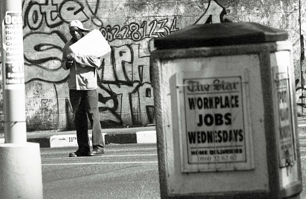 A man stands in an urban environment holding a large paper or cardboard object. The background features a graffiti-covered wall with bold lettering and numbers. In the foreground, an advertisement board displays a sign reading 'WORKPLACE JOBS WEDNESDAYS' in large letters. The scene is in black and white, creating a gritty, urban aesthetic.