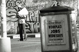 A man stands in an urban environment holding a large paper or cardboard object. The background features a graffiti-covered wall with bold lettering and numbers. In the foreground, an advertisement board displays a sign reading 'WORKPLACE JOBS WEDNESDAYS' in large letters. The scene is in black and white, creating a gritty, urban aesthetic.