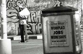 A man stands in an urban environment holding a large paper or cardboard object. The background features a graffiti-covered wall with bold lettering and numbers. In the foreground, an advertisement board displays a sign reading 'WORKPLACE JOBS WEDNESDAYS' in large letters. The scene is in black and white, creating a gritty, urban aesthetic.