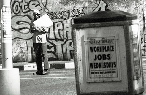 A man stands in an urban environment holding a large paper or cardboard object. The background features a graffiti-covered wall with bold lettering and numbers. In the foreground, an advertisement board displays a sign reading 'WORKPLACE JOBS WEDNESDAYS' in large letters. The scene is in black and white, creating a gritty, urban aesthetic.