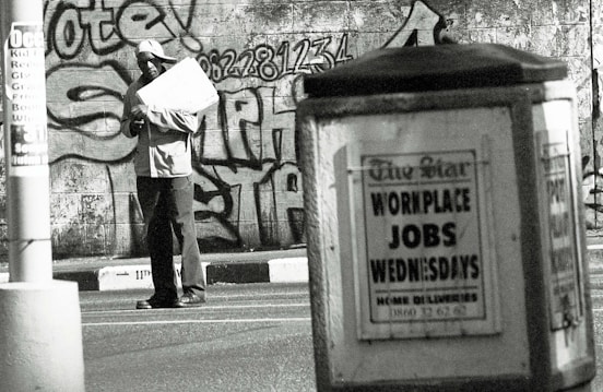 A man stands in an urban environment holding a large paper or cardboard object. The background features a graffiti-covered wall with bold lettering and numbers. In the foreground, an advertisement board displays a sign reading 'WORKPLACE JOBS WEDNESDAYS' in large letters. The scene is in black and white, creating a gritty, urban aesthetic.