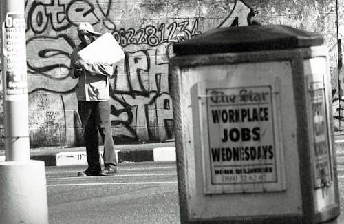 A man stands in an urban environment holding a large paper or cardboard object. The background features a graffiti-covered wall with bold lettering and numbers. In the foreground, an advertisement board displays a sign reading 'WORKPLACE JOBS WEDNESDAYS' in large letters. The scene is in black and white, creating a gritty, urban aesthetic.