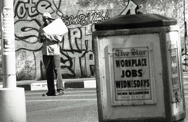 A man stands in an urban environment holding a large paper or cardboard object. The background features a graffiti-covered wall with bold lettering and numbers. In the foreground, an advertisement board displays a sign reading 'WORKPLACE JOBS WEDNESDAYS' in large letters. The scene is in black and white, creating a gritty, urban aesthetic.