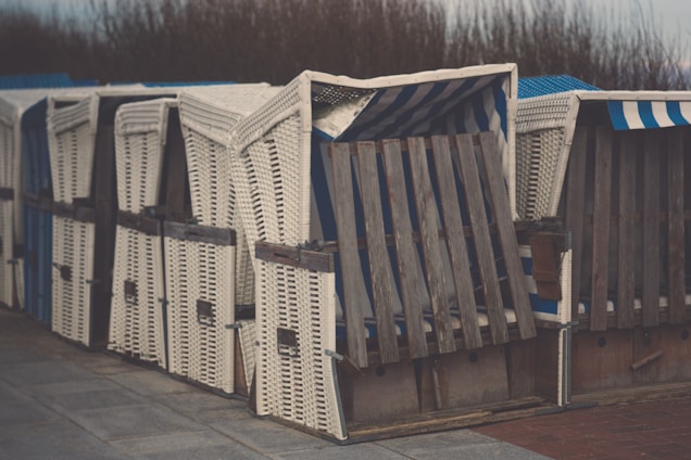 Several wicker beach chairs are aligned in a row, each with wooden panels and striped fabric inside. The chairs have a rustic, weathered appearance, and are situated outdoors on a paved area with some vegetation visible in the background.